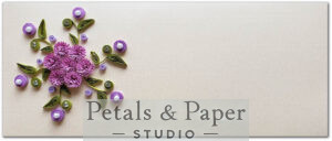 Handcrafted envelope featuring purple quilled flowers, green leaves, pearl accents, and gemstones on a beige textured background, ideal for gifting and special occasions.
