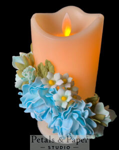 Beautiful LED candle with flickering flame, decorated with handcrafted blue and white flowers and green leaves, ideal for weddings, home décor, and gifting.
