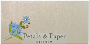 Handcrafted envelope with blue quilled flowers, green leaves, and rhinestone accents arranged in a paper cone on an ivory shimmer background, perfect for weddings, birthdays, and gifting occasions.