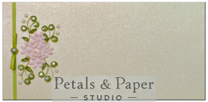 Handmade gift envelope with soft pink quilled flowers, green leaves, rhinestone and pearl accents, and a green ribbon bow on a shimmering cream background, perfect for gifting on special occasions.