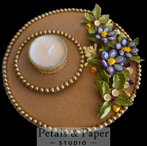 Handcrafted tea light holder plate with purple quilled flowers, green foliage, gold bead edging, and a gold-rimmed white candle for festive and wedding décor.