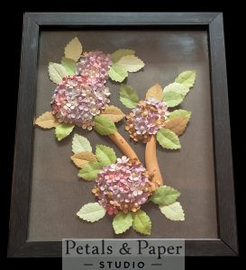 Handcrafted floral shadow box frame with pink and lavender paper flowers, green leaves, and wooden branch design in black frame for elegant home wall décor.